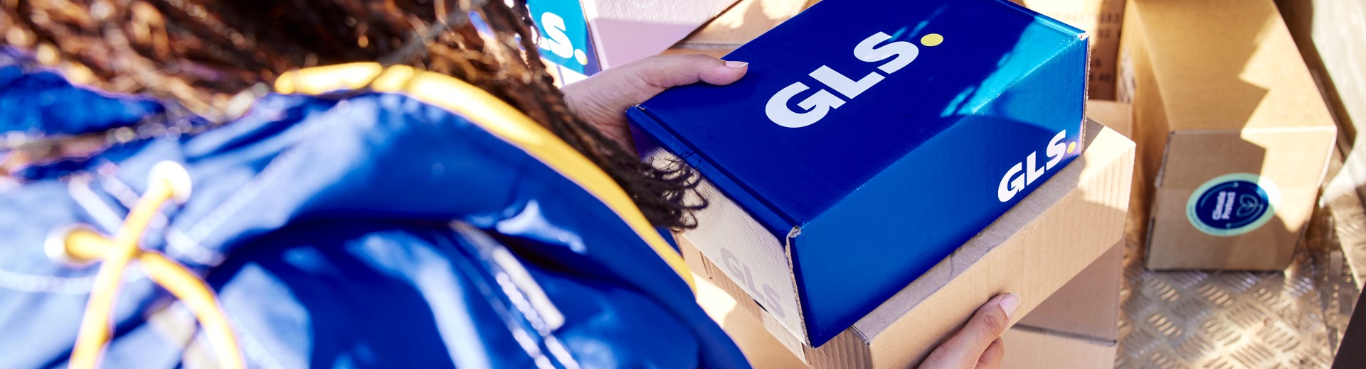 GLS driver photographed from behind holding GLS parcels in her hands, and the parcels are on board the GLS van