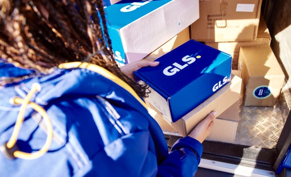 GLS driver photographed from behind holding GLS parcels in her hands, and the parcels are on board the GLS van