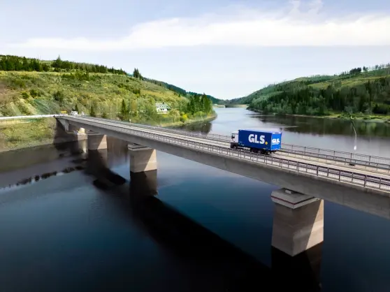 Camion bleu GLS sur un pont, dans un paysage naturel avec cours d'eau et végétation.