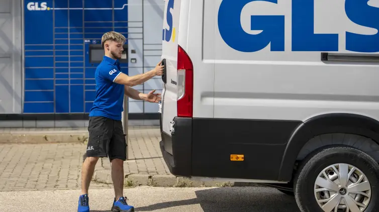 Livreur GLS en polo bleu ouvre la porte arrière d'un utilitaire GLS blanc, devant des consignes à colis GLS.