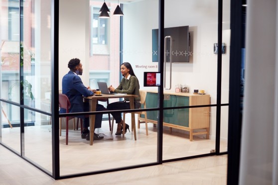 Deux professionnels en réunion dans une salle de meeting vitrée moderne, travaillant sur un ordinateur portable autour d’une table, illustrant la collaboration en entreprise et l’environnement de bureau.