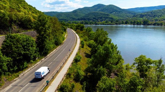 Une camionnette de livraison GLS circule sur une route sinueuse longeant un large fleuve, entourée d'une végétation luxuriante et de collines boisées. Le paysage verdoyant s'étend sous un ciel partiellement nuageux.