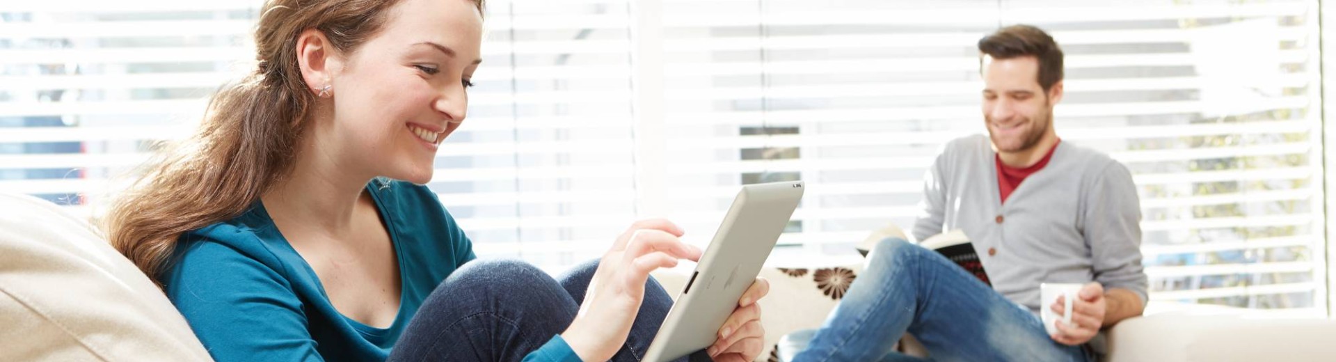 A woman using a tablet and a man reading a book while sitting on a couch.