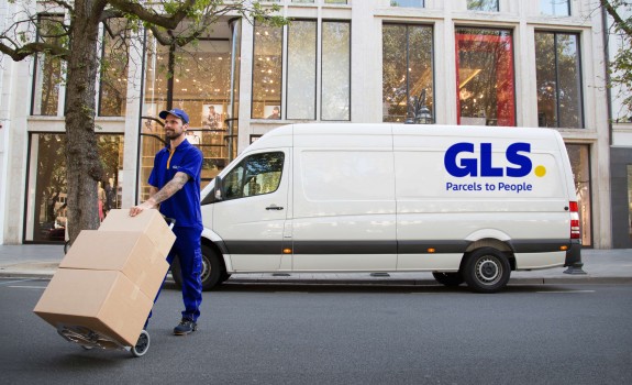 A GLS driver crosses the street, moving packages on a dolly in front of a GLS van.