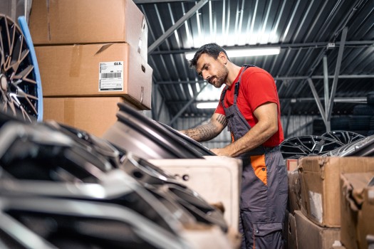 A warehouse worker inspects automotive parts beside stacked shipping boxes.